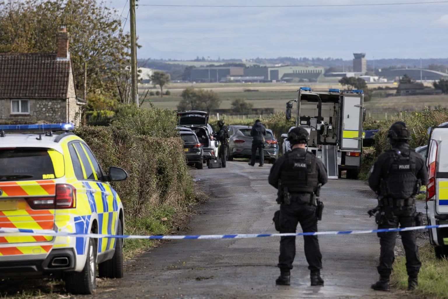 Police cordon on a rural village lane with police vehicles and an Army bomb disposal truck parked beyond the tape