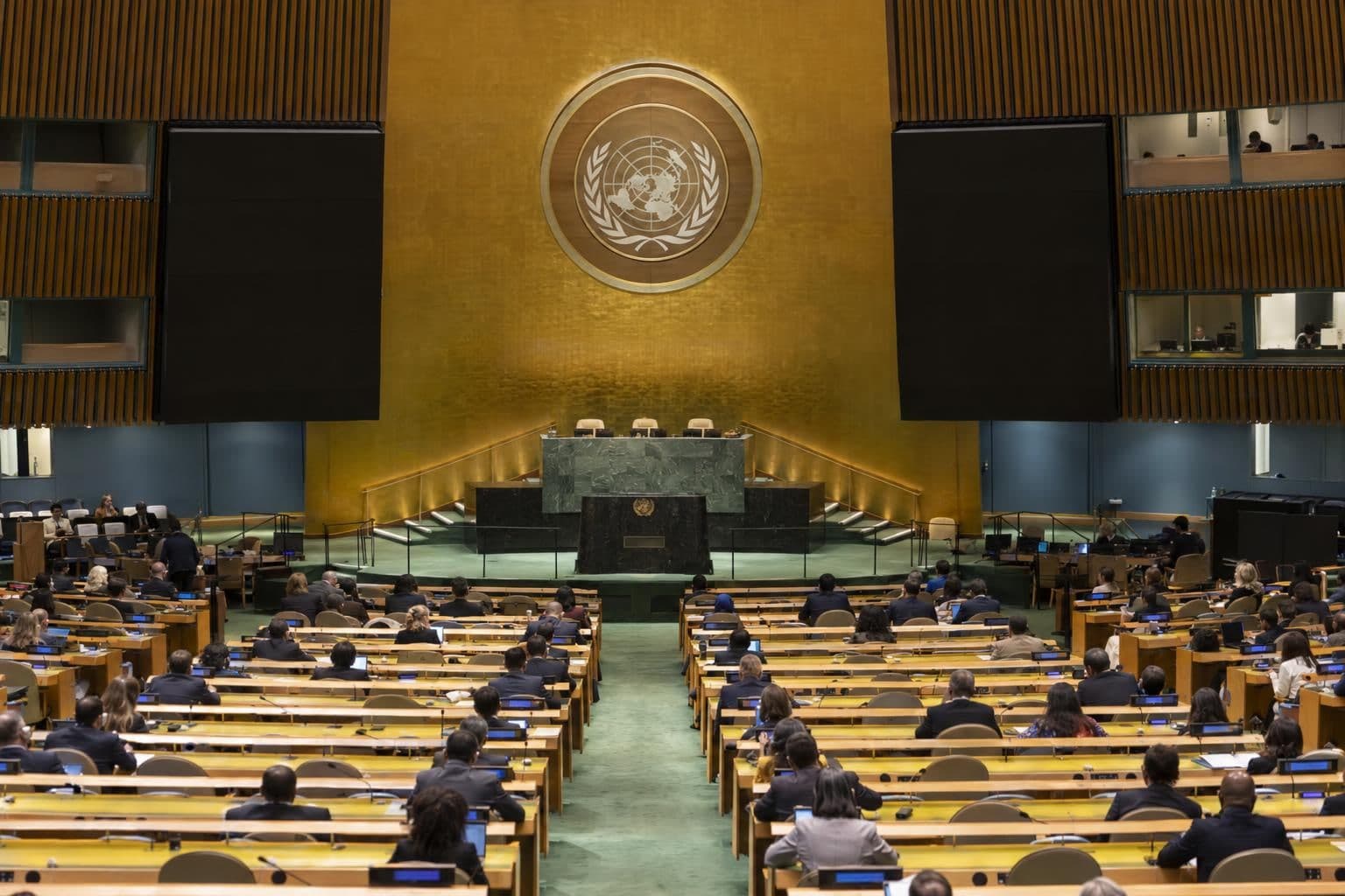 Delegates seated at curved desks facing an empty speaker's rostrum in the UN General Assembly hall