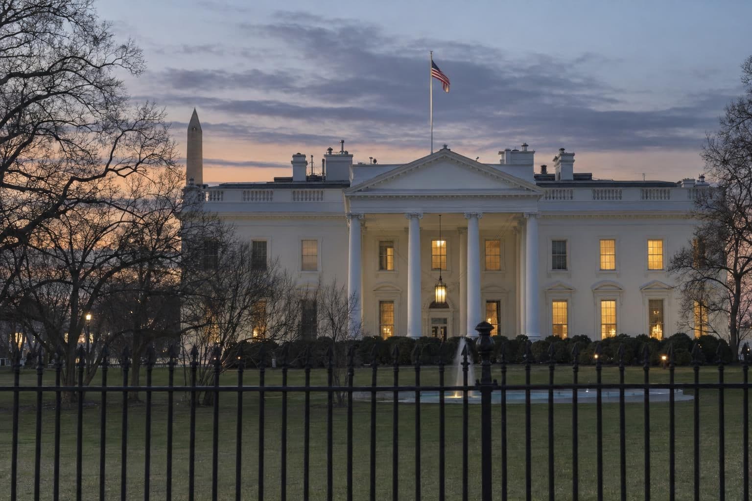 Exterior of the White House in Washington at evening, with light glowing in windows and an empty lawn in front.