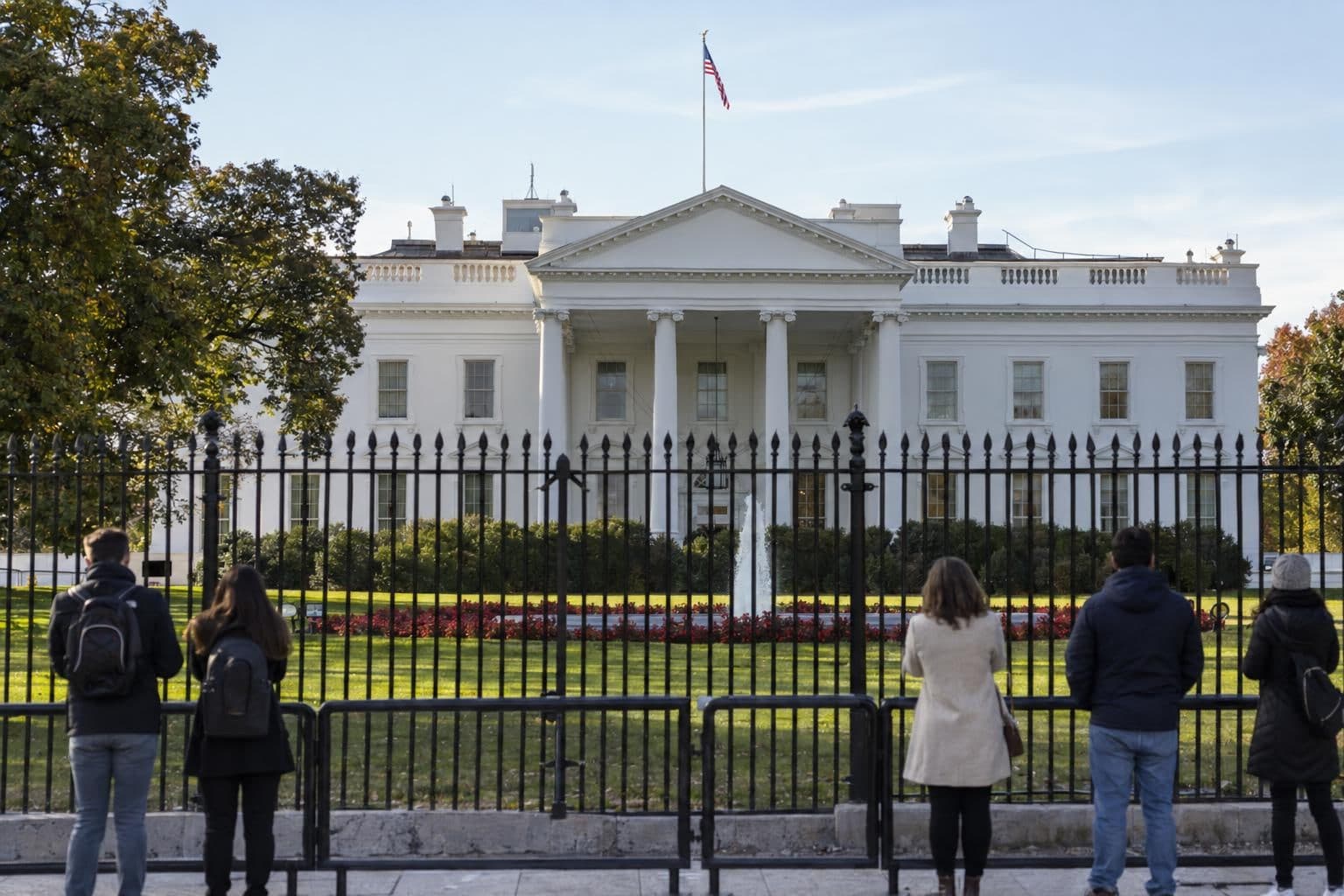 The White House north facade seen through its black iron fence, with a lawn and trees in front