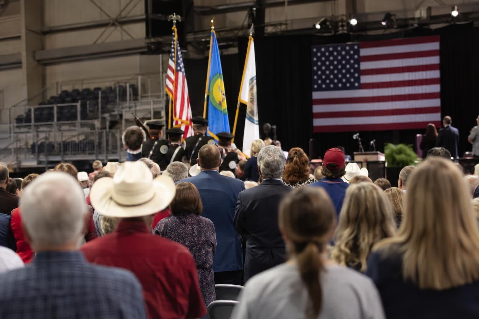 A large crowd gathers as a color guard presents flags at an indoor campaign rally.