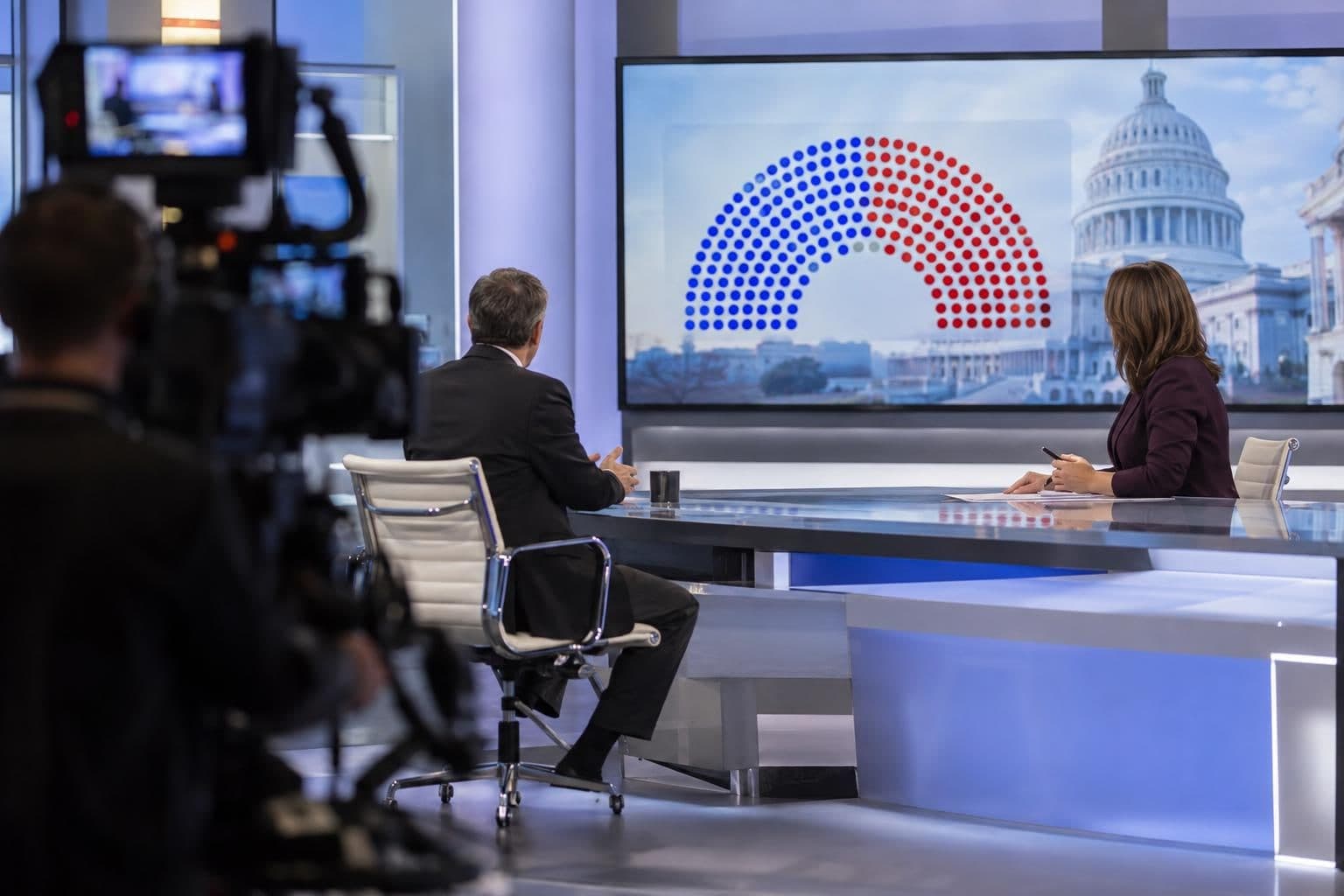 A political news studio shows a presenter beside a screen with a projected House map.
