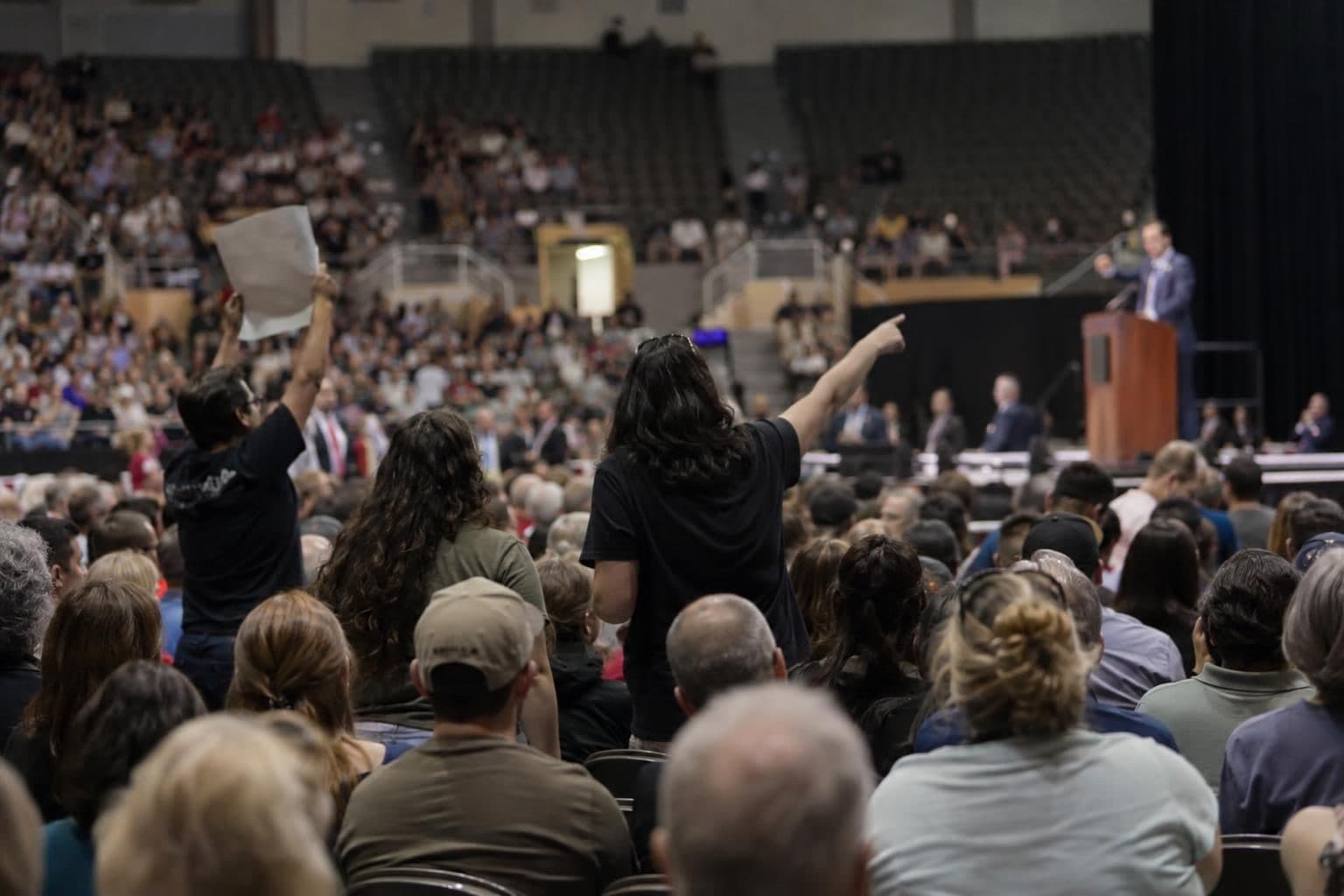 A speaker addresses a crowd at a campaign rally as protesters interrupt inside a coliseum.
