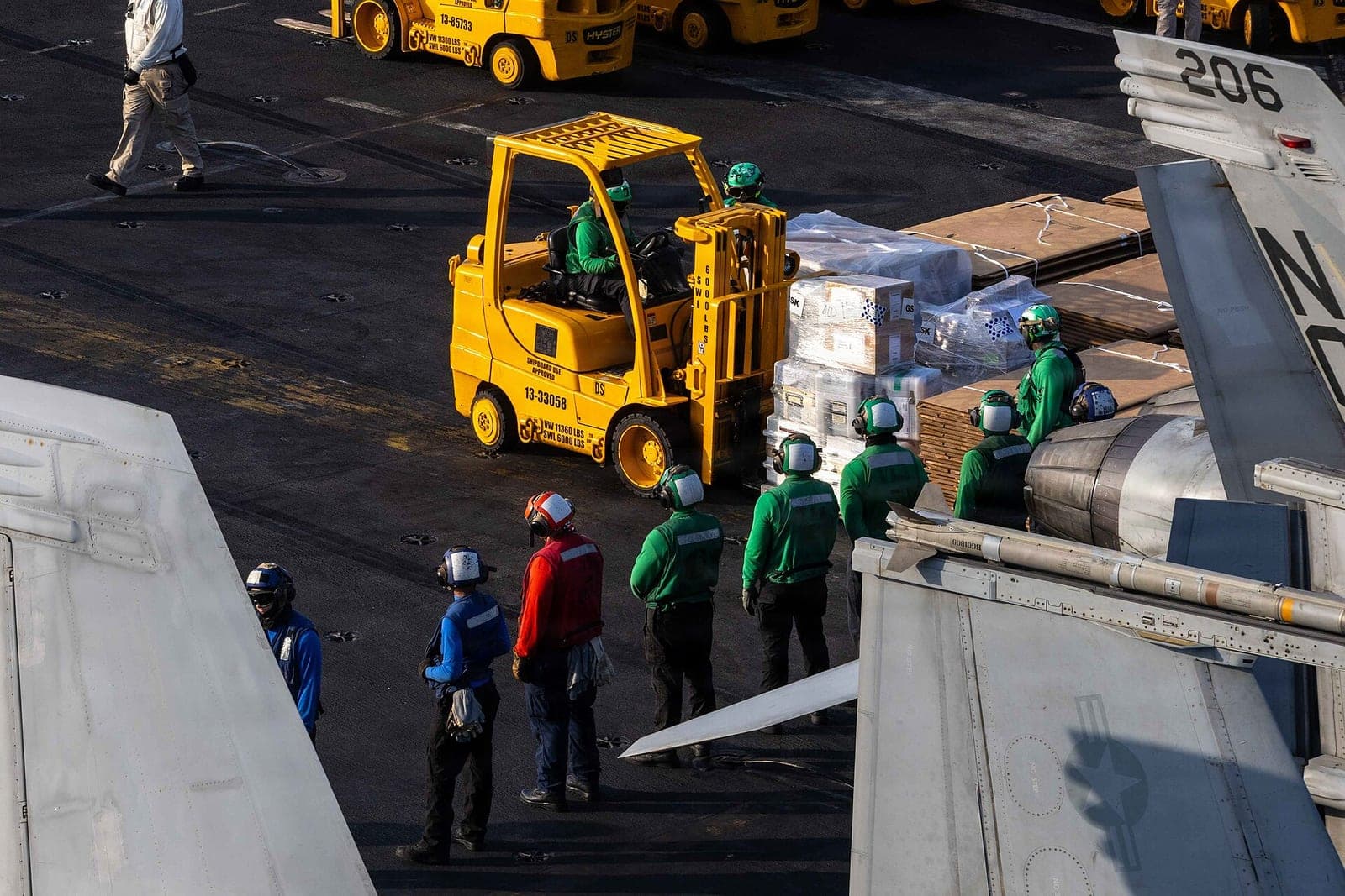 Sailors gather around a yellow forklift carrying palletized cargo on an aircraft carrier flight deck.