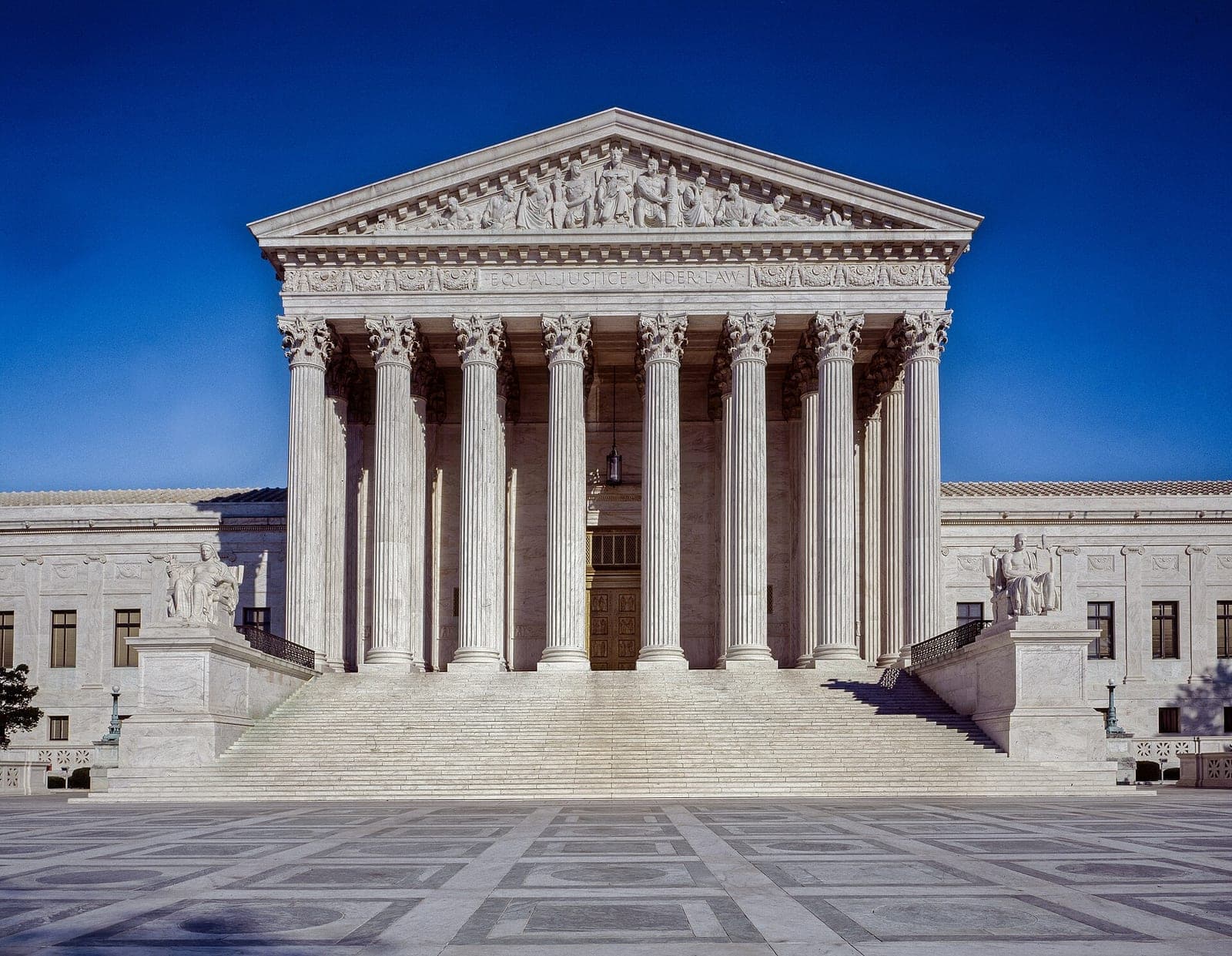 The U.S. Supreme Court building's columns and broad steps rise above an empty paved plaza under a blue sky.