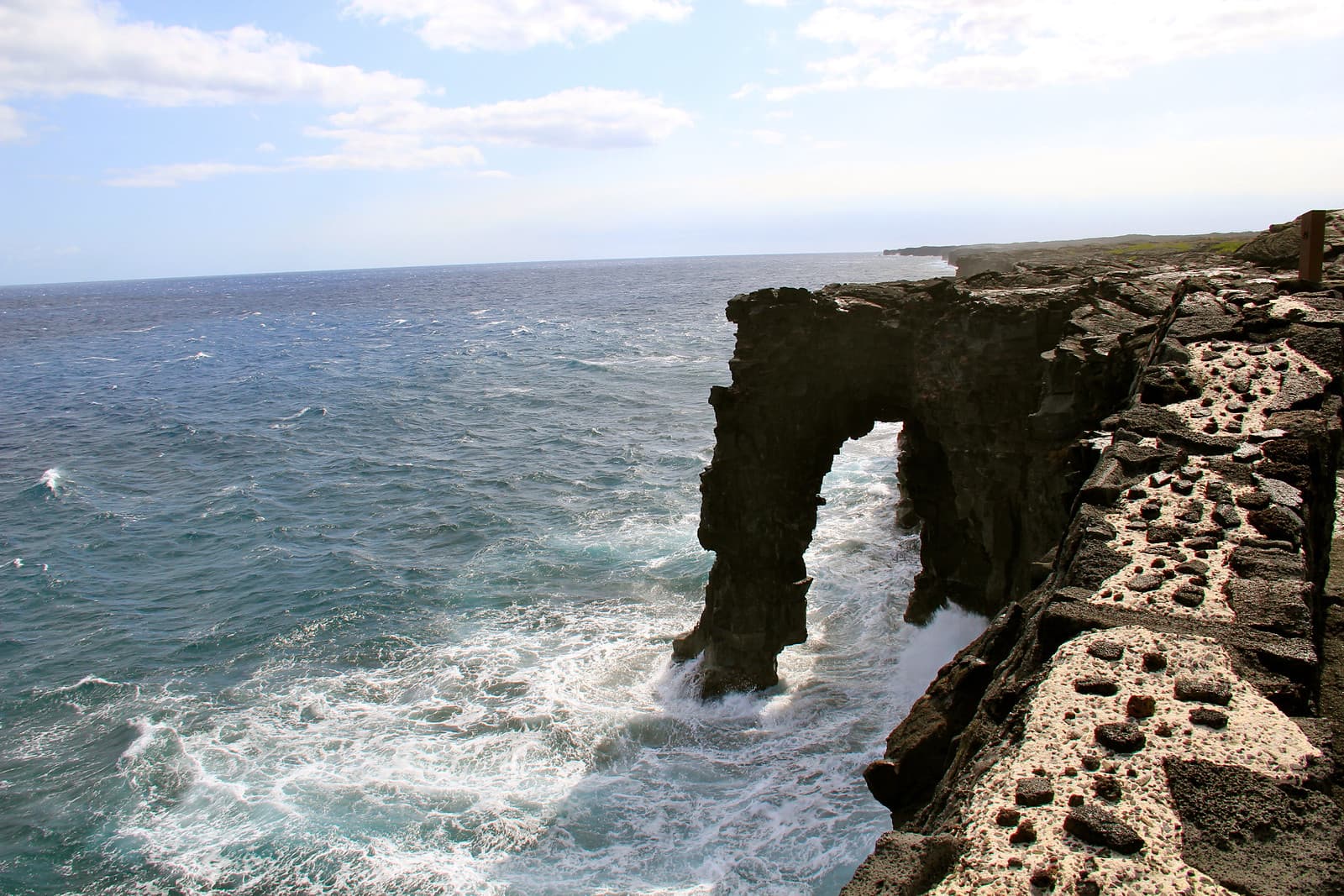 A dark rock arch extends over choppy ocean water beside a rugged coastal cliff.