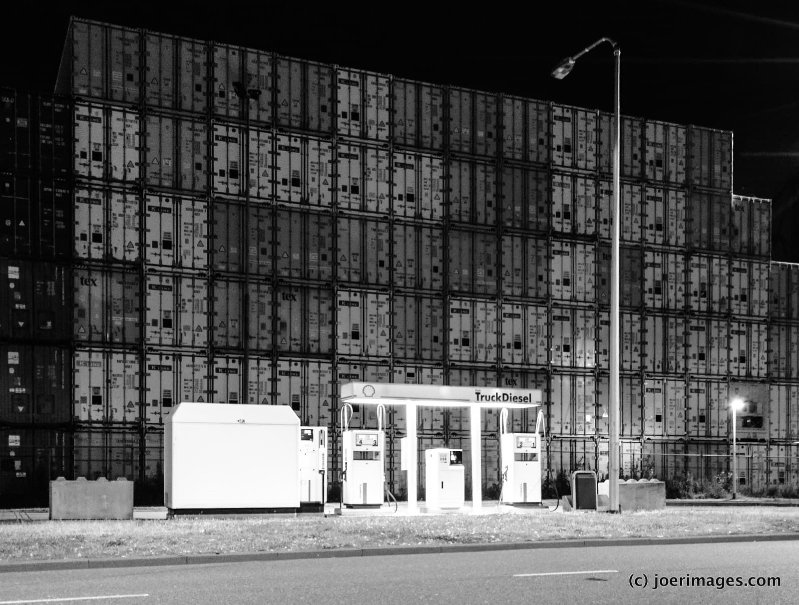 A diesel fueling station stands in front of stacked shipping containers.